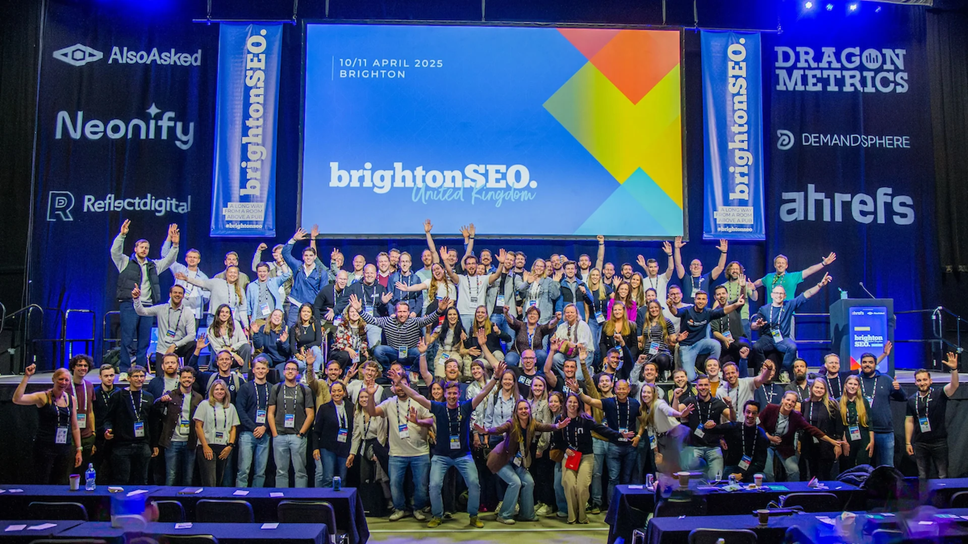 Foto grupal del cierre de BrightonSEO sobre el escenario, con asistentes levantando los brazos frente a banners de patrocinadores como Ahrefs, AlsoAsked y Dragon Metrics — guía de conferencias de SEO 2026